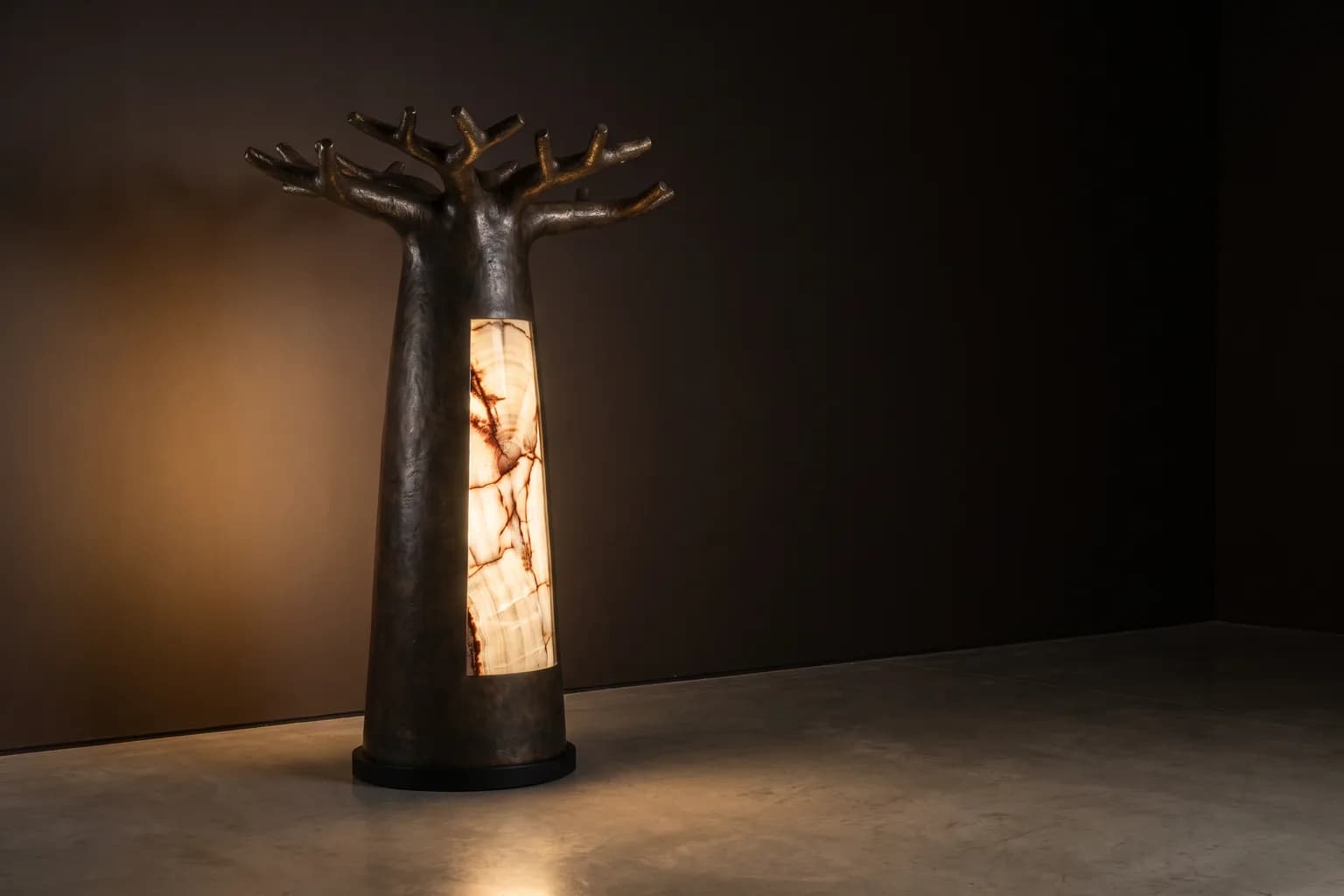 Visualisation éditoriale d'un lampadaire en bronze sombre en forme de baobab, dont le tronc porte un insert de bois pétrifié rétroéclairé